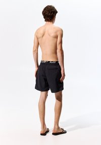 Pantaloni da bagno neri con cintura elastica e dettaglio del logo bianco, tessuto leggero, lunghezza sopra il ginocchio, abbinati a ciabatte nere.