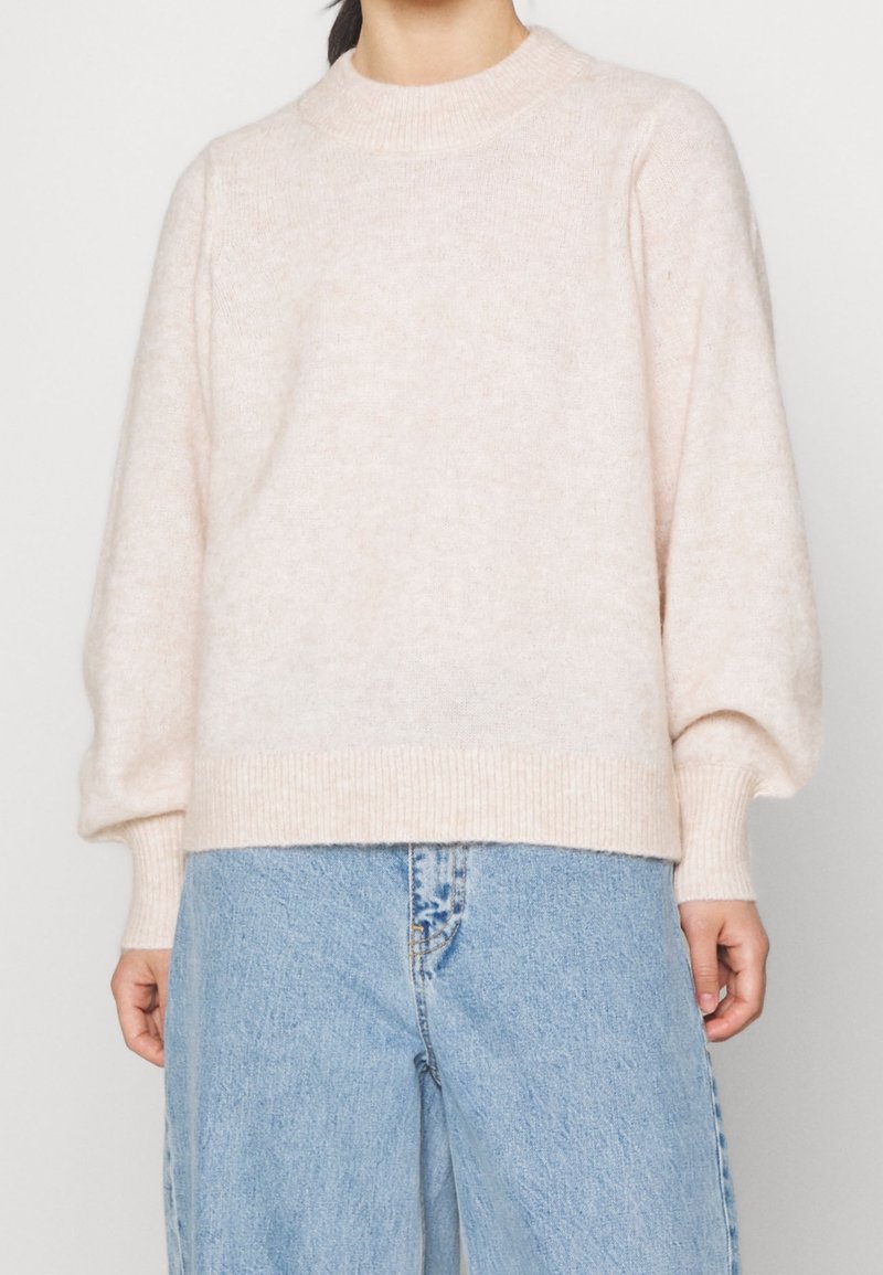 Pull en tricot texturé rose clair avec col montant, épaules tombantes et poignets côtelés, associé à un jean bleu clair taille haute.