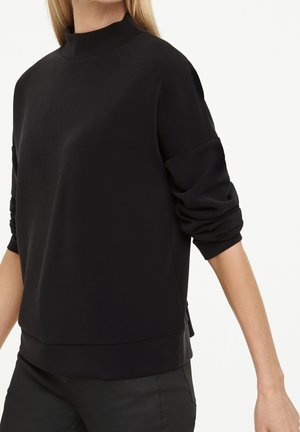Femme portant un pull noir à manches longues avec un col montant et des manches retroussées, associé à un pantalon noir.