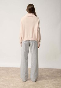 Przesadnie duży jasnoróżowy sweter z wysokim kołnierzem, w połączeniu z szerokimi spodniami dżinsowymi w odcieniu szarości. Miękka tekstura i luźny krój to charakterystyczne cechy.