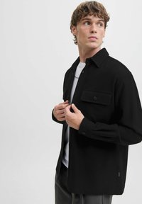 Camicia nera con colletto, maniche lunghe, tasca sul petto anteriore e vestibilità comoda. Indossata sopra una camicia bianca, texture liscia.