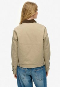 Veste bomber beige avec un col en velours côtelé marron, tissu texturé, poches zippées et coupe ajustée, associée à un jean en denim bleu.