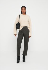 Pullover di crema morbido, pantaloni grigi a vita alta con texture e stivaletti neri alla caviglia. Modella che tiene una piccola borsa nera, in piedi contro uno sfondo bianco.
