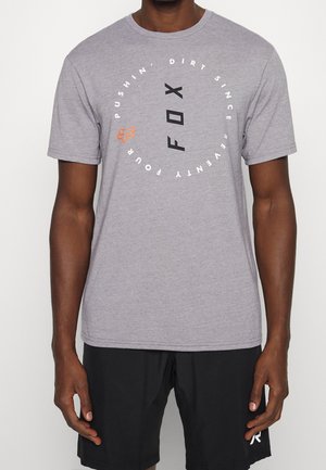 Eine Person trägt ein graues T-Shirt mit einem kreisförmigen Grafikdesign und Text, steht vor einem schlichten Hintergrund und trägt schwarze Shorts.