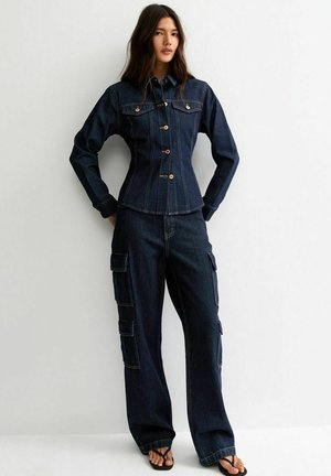 Vrouw draagt een donker denim jack met knopen en wijde cargobroek, staand tegen een effen lichte achtergrond.