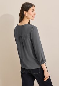 Blusa gris de manga larga con escote redondeado, detalle de pliegues en la parte posterior y puños elásticos, combinada con jeans oscuros frente a un fondo neutro.