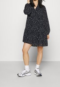 Abito nero con macchie bianche, maniche lunghe e scollatura a V. Indossato con scarpe sportive argentate e calze bianche.