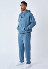Sudadera de algodón azul con bolsillo delantero y cordón, combinada con pantalones de chándal a juego. Zapatillas blancas completan el atuendo.
