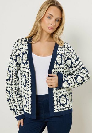 Blonde Frau, die eine marineblaue und weiße gemusterte Strickjacke über einem weißen Oberteil und dunklen Hosen trägt, stehend vor einem einfarbigen Hintergrund.