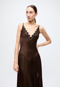 Brown satin long dress with black lace trim, deep V-neckline, thin straps, and a pleated skirt featuring a side slit.