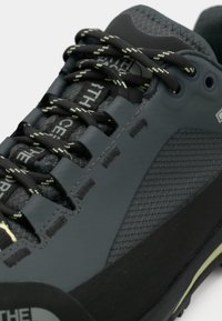 Fekete sportcipő texturált felülettel, szürke részletekkel és kontrasztos zöld fűzőkkel. Gumi talp megerősített orrával.