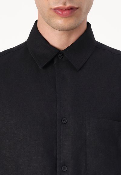 Chemise noire à boutons en tissu texturé avec un col pointu, dotée d'une poche poitrine unique et de boutons noirs.