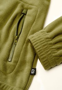 Tissu polaire vert olive avec une poche zippée, un poignet élastique et une petite étiquette noire avec un logo blanc.
