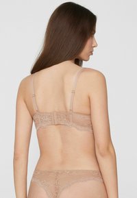 Kanten beha in naaktkleur met verstelbare bandjes, haaksluiting en bloemmotiefdetails langs de achterkant en band.