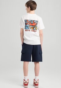 White cotton t-shirt featuring a colorful Keith Haring design on the back, paired with navy shorts that have two side pockets.
