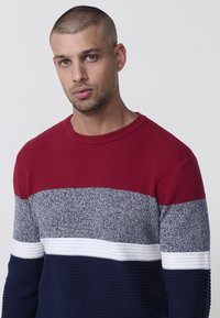 Maglione a righe rosso, grigio e blu navy con una texture a coste. Design a collo rotondo con bande orizzontali a contrasto e una striscia bianca liscia.