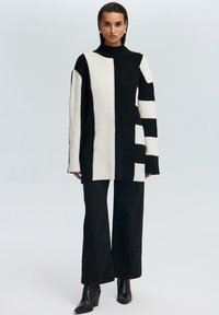 Zwart-witte geribbelde coltrui met asymmetrisch blokpatroon; gecombineerd met zwarte wijde broek en enkellaarsjes.