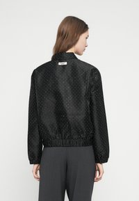 Čierna saténová bunda s bodkovaným vzorom, elastickým pásom a mierne nafúknutými rukávmi. Na golieri je viditeľná štítok zo zadnej strany.