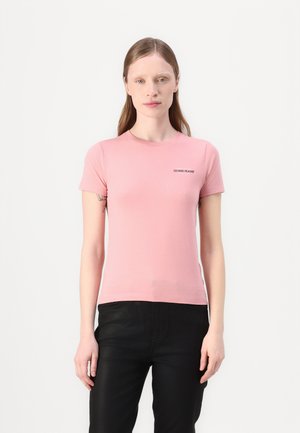Rosa T-Shirt mit kurzen Ärmeln aus weichem Material, runder Ausschnitt und schwarzem "Guess Jeans" Logo auf der linken Seite. Kombination mit schwarzen Hosen.