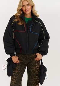 Čierna pérová bunda s farebnými lemami v červenej, modrej a zelenej, s vysokým golierom a elastickými manžetami, v kombinácii s nohavicami so vzorom leopardieho potlače.