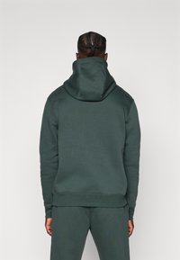 Uomo con capelli intrecciati che indossa un felpa verde scuro e pantaloni abbinati visti di spalle su uno sfondo neutro.