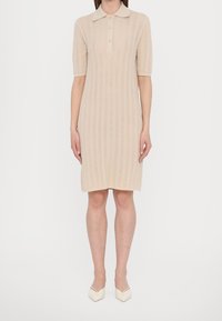 Femme portant une robe en maille beige longueur genou, manches courtes, col, boutons, et des chaussures beiges à bouts pointus, debout sur fond blanc.