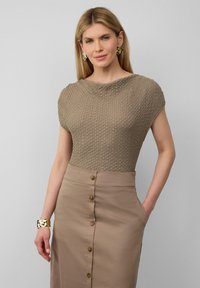 Beige strukturierter Oberteil mit kurzen Ärmeln, kombiniert mit einem hochtaillierten braunen Rock mit vorderen Knöpfen. Goldene Armreifenakzent am Handgelenk.