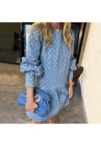 Femme portant une robe bleue à fleurs avec des manches bouffantes et tenant une pochette bleue assortie avec une boucle décorative.