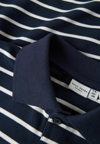 Marinblå polo skjorta med vita horisontella ränder, med krage, knappslå och vävd textur. Tvättrådsskylt synlig.