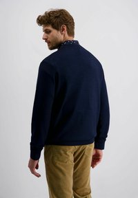 Marineblauer Pullover mit strukturierter Oberfläche und gemustertem Kragen, kombiniert mit hellbraunen Cordhosen. Das Modell ist von hinten zu sehen.
