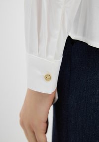 Camicia bianca con dettaglio a manica plissettata, caratterizzata da un bottone dorato al polsino. Indossata con denim scuro.