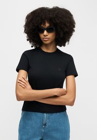 Γυναίκα με σγουρά μαλλιά που φοράει μαύρα γυαλιά ηλίου, εφαρμοστό μαύρο μπλουζάκι και μπλε τζιν ψηλόμεσο, με τα χέρια σταυρωμένα.