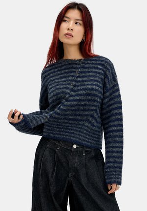 Jeune femme aux longs cheveux roux porte un pull boutonné rayé bleu marine et gris, en tissu duveteux, et un pantalon ample foncé sur fond blanc.