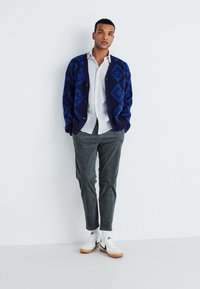 Blau- und schwarz gemusterte Strickjacke, weißes Hemd mit Knöpfen, graue Cordhosen und weiße Sneakers mit schwarzen Akzenten. Stehende Pose vor einer weißen Wand.