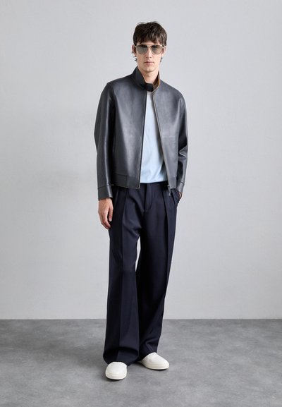 Schwarze Lederbomberjacke mit hohem Kragen, kombiniert mit einem hellblauen Hemd, maßgeschneiderte marineblaue Hose und weißen Sneakers.