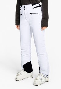 Person, der weiße Skihosen mit schwarzen Flecken an den Knöcheln und weiße Skischuhe trägt, steht vor einem weißen Hintergrund.