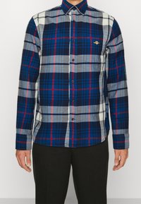 Camisa de cuadros en tela azul, gris y roja con mangas largas, cuello tipo botón y un pequeño logo bordado en el pecho.