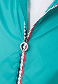 Tyrkijsblauwe lichtgewicht jas met een contrasterend rood-blauw ritssluitingdetail, een zilveren ronde ritssluitingtrekker en een gestructureerde stof.