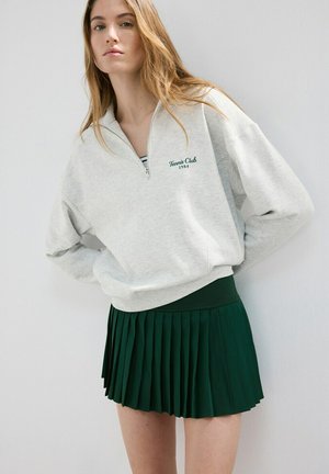Mujer joven con sudadera gris clara de cremallera hasta un cuarto, con la inscripción "Tennis Club 1984" y falda plisada verde oscuro, con fondo blanco liso.