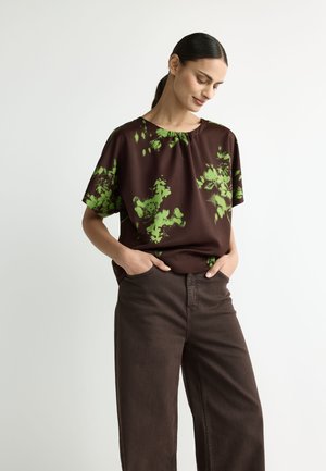 Woman with dark hair in a ponytail wearing a brown blouse with green floral patterns and dark brown pants, hands in pockets, looking down.