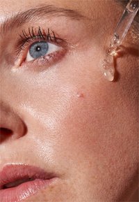 Nahaufnahme eines Frauengesichts mit blauen Augen und einem klaren Serumtröpfchen aus einem Pipettenbehälter in der Nähe der Wange.