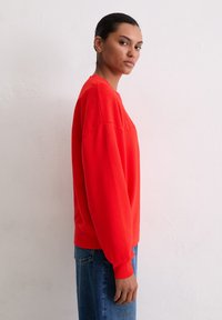 Czerwona bluza z miękkiego materiału, z okrągłym dekoltem i ściągaczami przy mankietach. Połączona z niebieskimi dżinsami, pokazana z boku.