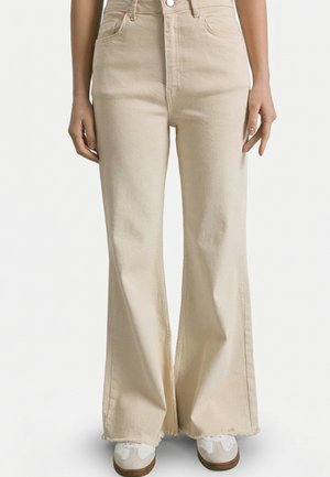 Beige vidde bukser med høj talje og flossede kanter, båret med hvide sneakers, vist fra talje til fødder på en neutral baggrund.