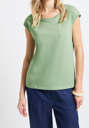 Vrouw draagt een lichtgroen T-shirt met korte mouwen, donkerblauwe spijkerbroek en een lichtgekleurde armband om haar rechterpols.
