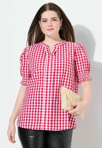 Chemise vichy rouge et blanche avec un col en V et des manches bouffantes. Portée avec un pantalon en cuir noir et tenant une pochette tissée.