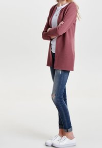 Kobieta stoi z rękami skrzyżowanymi, ubrana w białą koszulę, mauve kardigan, podarte niebieskie dżinsy i białe trampki na jednolitym białym tle.