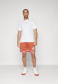 Conjunto masculino que incluye una camiseta de algodón blanca con un logo negro, pantalones cortos de malla naranja con detalles blancos y zapatillas blancas con acentos rojos.