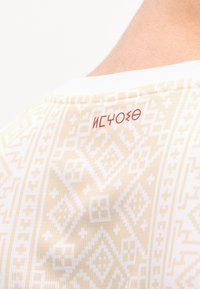 Ύφασμα σε μπεζ και λευκό με γεωμετρικά σχήματα και κόκκινο κείμενο κοντά στον λαιμό στο πίσω μέρος ενός ατόμου με ανοιχτόχρωμο δέρμα.