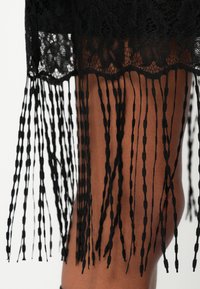 Melnas mežģīņu kleita ar viļņotu malu, kas rotāta ar garām bārkstīm no mezglotas dzijas, radot teksturētu un daudzslāņu izskatu.