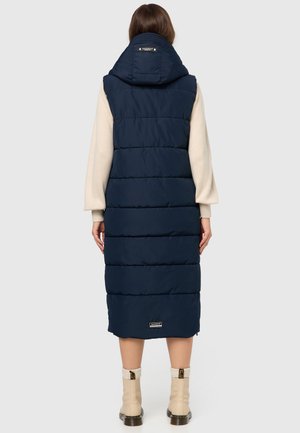 Vesta lungă de puf de culoare bleumarin, cu glugă, design matlasat și fermoar frontal. Însoțită de cizme la glezna de culoare bej, cu accente texturate.